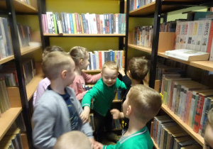 Dzieci zwiedzają bibliotekę, oglądają książki na regałach.
